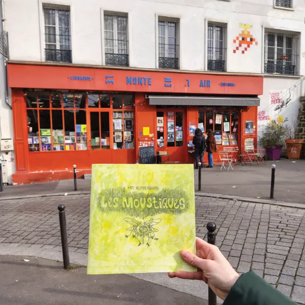 In the foreground: a hand holding a print version of the comic. In the background : a bookshop