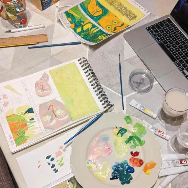 A table with sketches and a work in progress of a gouache painting of the comic